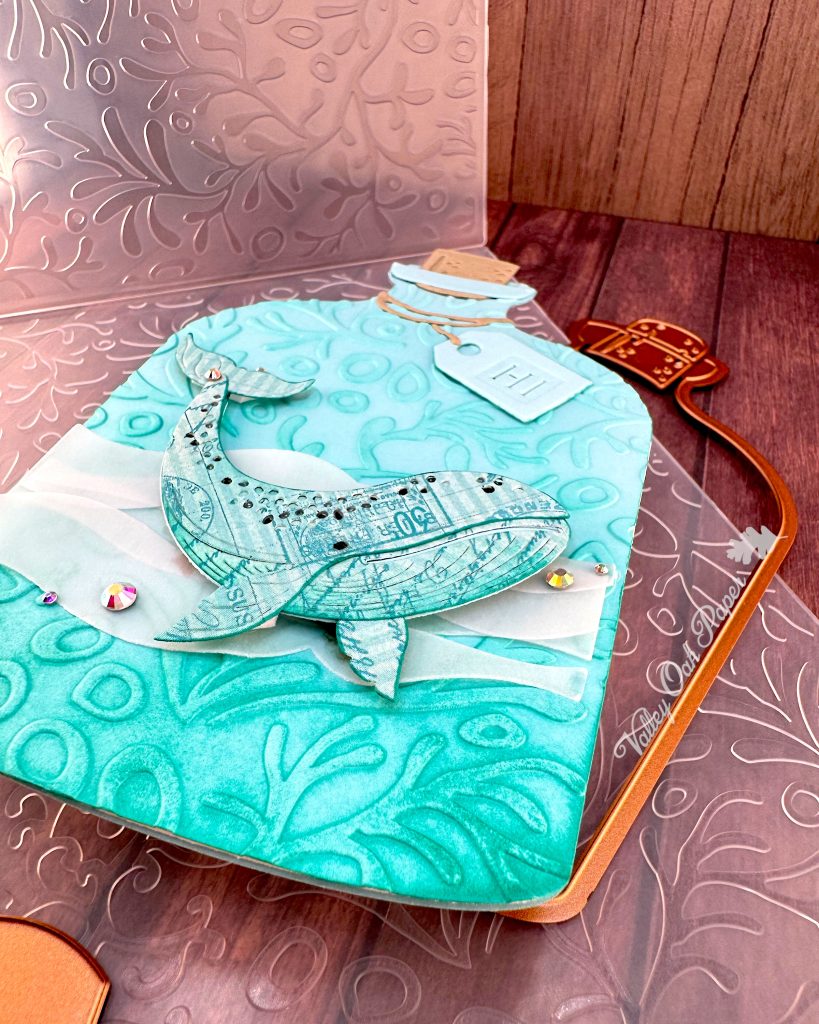 A handmade greeting card in the shape of a turquoise glass bottle with a cork stopper and "Hi" tag. The bottle contains a humpback whale swimming in stylized vellum ocean waves. The bottle has been embossed with a coral pattern.
The card is sitting above the coral pattern embossing folder used to create it. Under it some of the dies used when creating it are peeking through. It is pictured on a wooden surface. The Valley Oak Paper logo is visible on the right edge of the bottle die.