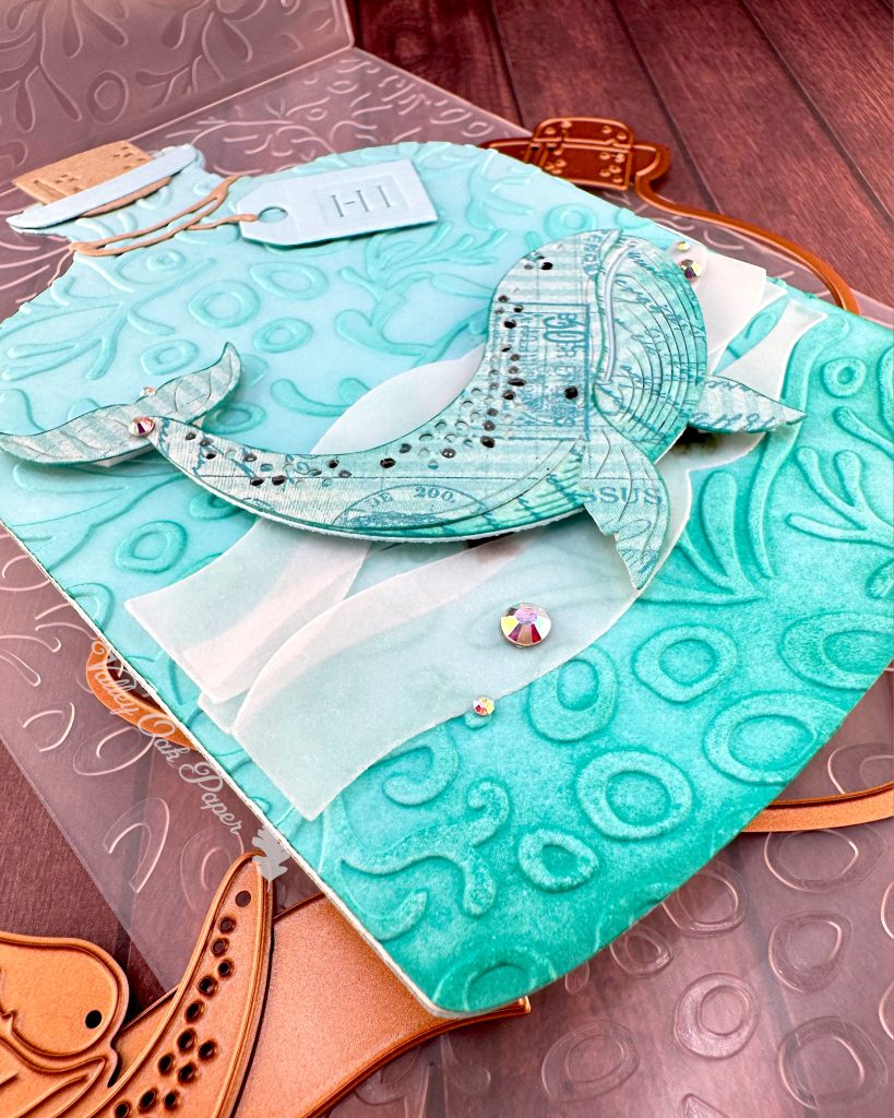 A handmade greeting card in the shape of a turquoise glass bottle with a cork stopper and "Hi" tag. The bottle contains a humpback whale swimming in stylized vellum ocean waves. The bottle has been embossed with a coral pattern.
The card is sitting above the coral pattern embossing folder used to create it. Under it some of the dies used when creating it are peeking through. It is pictured on a wooden surface. The Valley Oak Paper logo is visible on the right edge of the card.