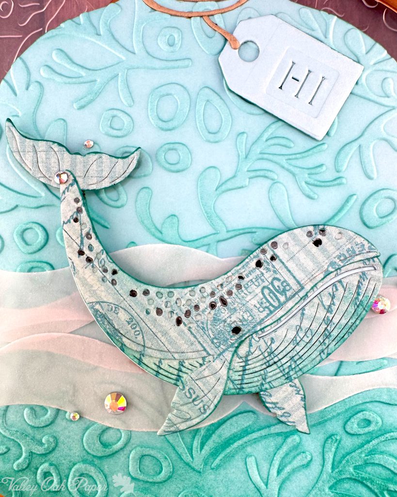 A closeup of a handmade greeting card in the shape of a turquoise glass bottle with a "Hi" tag. The bottle contains a humpback whale swimming in stylized vellum ocean waves. The bottle has been embossed with a coral pattern.
The Valley Oak Paper logo is visible in the bottom left corner.
