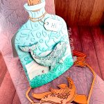 A handmade greeting card in the shape of a turquoise glass bottle with a cork stopper and "Hi" tag. The bottle contains a humpback whale swimming in stylized vellum ocean waves. The bottle has been embossed with a coral pattern. In front of it are some of the dies used when creating it. The card stands on the coral pattern embossing folder used to create it. It is pictured on a wooden surface. In the top center, the Valley Oak Paper logo is visible.