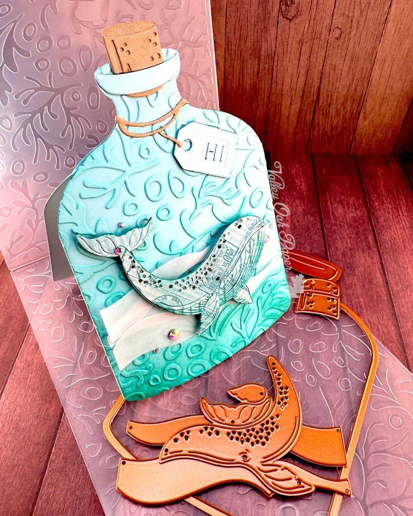 A handmade greeting card in the shape of a turquoise glass bottle with a cork stopper and "Hi" tag. The bottle contains a humpback whale swimming in stylized vellum ocean waves. The bottle has been embossed with a coral pattern.
In front of it are some of the dies used when creating it. The card stands on the coral pattern embossing folder used to create it. It is pictured on a wooden surface. The Valley Oak Paper logo is visible on the right edge of the bottle-shaped card.