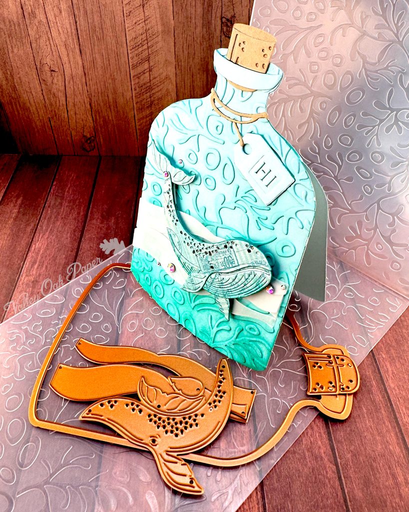 A handmade greeting card in the shape of a turquoise glass bottle with a cork stopper and "Hi" tag. The bottle contains a humpback whale swimming in stylized vellum ocean waves. The bottle has been embossed with a coral pattern.
The card stands on the coral pattern embossing folder used to create it. In front of it are some of the dies used when creating it. It is pictured on a wooden surface. The Valley Oak Paper logo is visible on the left edge of the embossing folder.