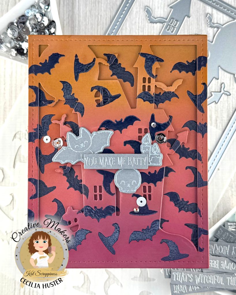 A handmade Halloween greeting card featuring an orange to dark pink gradient background filled with dark purple heat embossed bat silhouettes. The outline of a haunted mansion can also be seen.
The card has a decorative stitched border and contains silver embellished elements including a banner that reads "YOU MAKE ME BATTY." Around the sentiment a bat, a skull and some candles are grouped.
The card is photographed on a white surface alongside metal cutting dies, sequins in a triangle tray and other cardmaking supplies.
In the bottom left corner is a circular logo for "Creative Makers" featuring a cartoon woman with brown hair, credited to "Kat Scrappiness" and "CECILIA HUSTER."