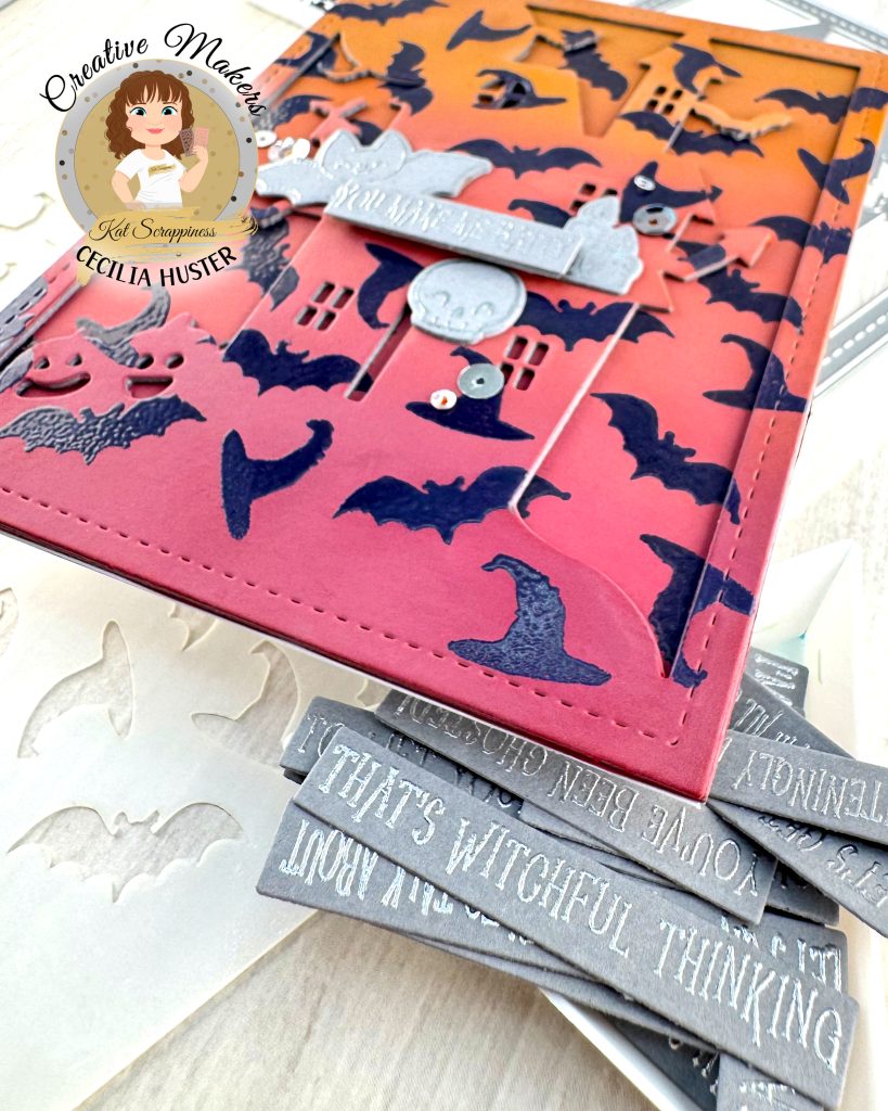 A handmade Halloween greeting card featuring an orange to dark pink gradient background filled with dark purple heat embossed bat silhouettes. The outline of a haunted mansion can also be seen.
The card has a decorative stitched border and contains silver embellished elements including a banner that reads "YOU MAKE ME BATTY." Around the sentiment a bat, a skull and some candles are grouped.
The card is photographed lying on a white surface alongside more silver and gray Halloween sentiment banners in a triangle tray, a stencil and other cardmaking supplies.
In the top left corner is a circular logo for "Creative Makers" featuring a cartoon woman with brown hair, credited to "Kat Scrappiness" and "CECILIA HUSTER."