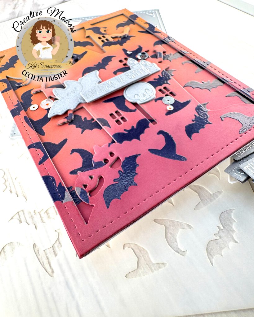 A handmade Halloween greeting card featuring an orange to dark pink gradient background filled with dark purple heat embossed bat silhouettes. The outline of a haunted mansion can also be seen.
The card has a decorative stitched border and contains silver embellished elements including a banner that reads "YOU MAKE ME BATTY." Around the sentiment a bat, a skull and some candles are grouped.
The card is photographed lying on a white surface alongside metal craft dies, a stencil and other cardmaking supplies.
In the top left corner is a circular logo for "Creative Makers" featuring a cartoon woman with brown hair, credited to "Kat Scrappiness" and "CECILIA HUSTER."