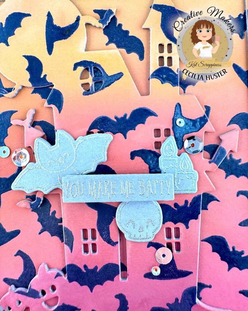 A close-up of a handmade Halloween greeting card featuring an orange to dark pink gradient background filled with dark purple heat embossed bat silhouettes. The outline of a haunted mansion can also be seen.
The focus of the card is a banner that reads "YOU MAKE ME BATTY." Around the sentiment a bat, a skull, some candles and silver sequins are grouped.
In the top right corner is a circular logo for "Creative Makers" featuring a cartoon woman with brown hair, credited to "Kat Scrappiness" and "CECILIA HUSTER."