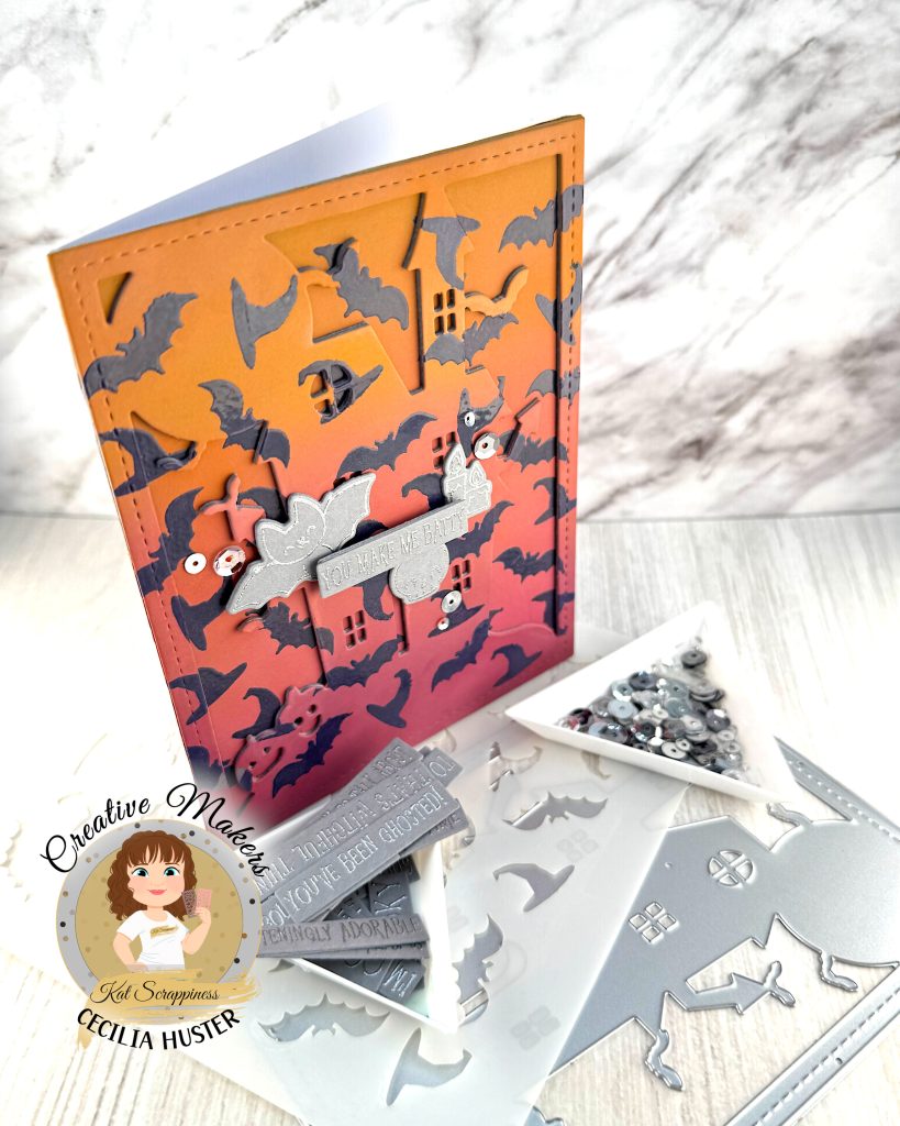 A handmade Halloween greeting card featuring an orange to dark pink gradient background filled with dark purple heat embossed bat silhouettes. The outline of a haunted mansion can also be seen.
The card has a decorative stitched border and contains silver embellished elements including a banner that reads "YOU MAKE ME BATTY." Around the sentiment a bat, a skull and some candles are grouped.
The card is photographed standing up and facing right on a white surface alongside metal cutting dies, sequins in a triangle tray, a stencil and other cardmaking supplies.
In the bottom left corner is a circular logo for "Creative Makers" featuring a cartoon woman with brown hair, credited to "Kat Scrappiness" and "CECILIA HUSTER."
