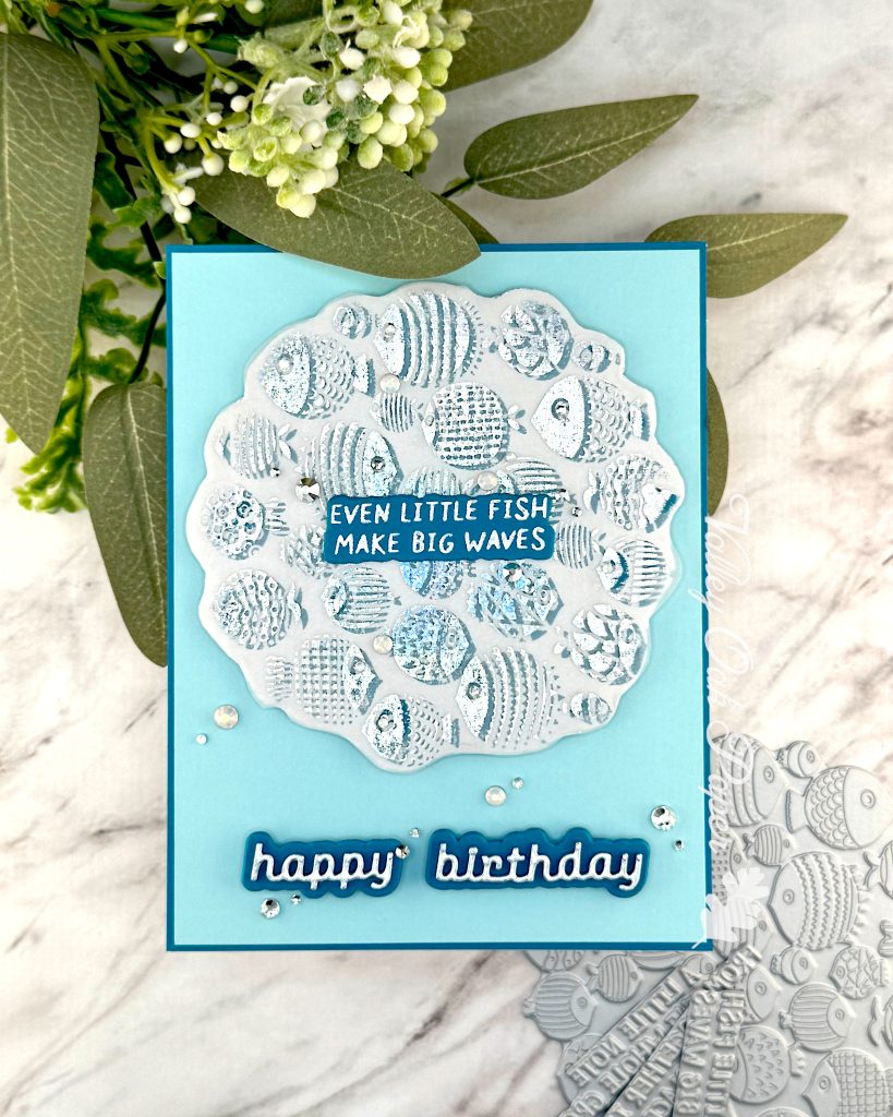 A handmade birthday card with a light turquoise background featuring a scalloped vellum circle filled with silver foiled fish designs in various patterns and textures. In the center of the fish pattern is a dark turquoise sentiment with silver text reading "EVEN LITTLE FISH MAKE BIG WAVES." 

Below the circular design, "happy birthday" appears in silver cursive lettering on a dark turquoise shadow layer. 

Small clear rhinestones or gems are scattered across the card for decoration. The card is photographed on a white marble surface with green foliage and small flowers visible in the upper portion of the image, and part of a BetterPress plate with fish designs is visible in the bottom right corner.