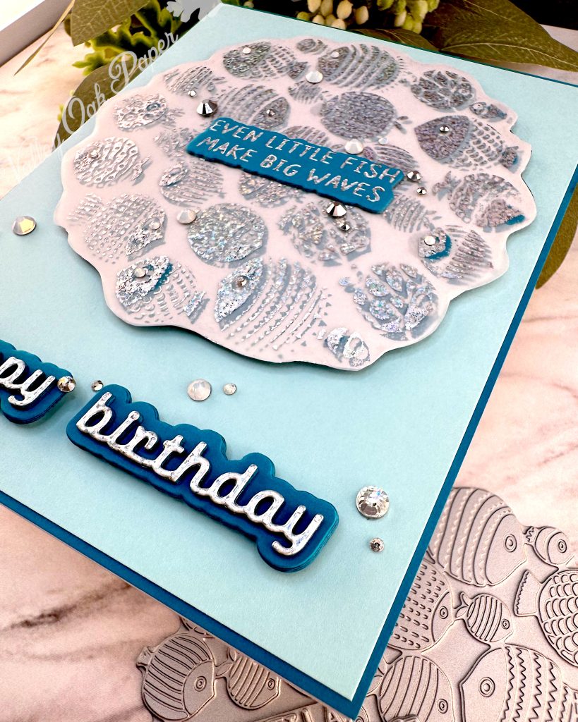 A handmade birthday card with a light turquoise background featuring a scalloped vellum circle filled with silver foiled fish designs in various patterns and textures. In the center of the fish pattern is a dark turquoise sentiment with silver foiled text reading "EVEN LITTLE FISH MAKE BIG WAVES." 

Below the circular design, "happy birthday" appears in silver holographic lettering on a dark turquoise shadow layer. 

Small clear rhinestones or gems are scattered across the card for decoration. The card is photographed on a white marble surface with green foliage and small flowers visible in the upper portion of the image and a press plate with fish in the lower right corner. The card is photographed from the bottom right. 