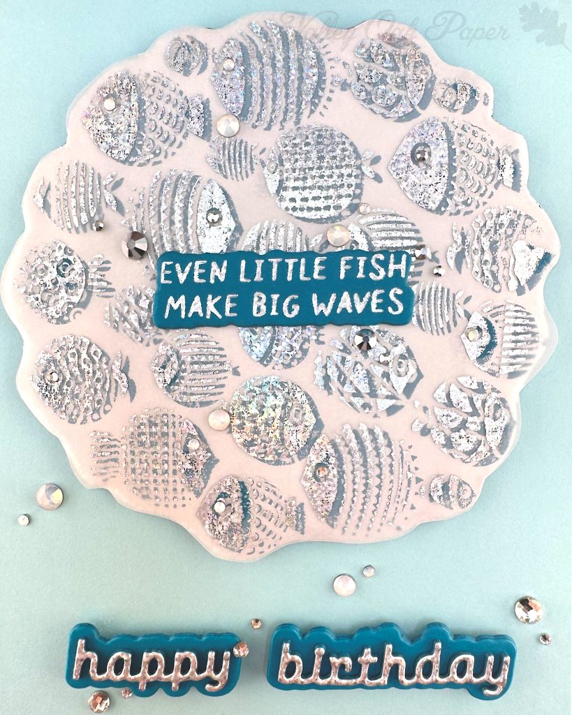 Closeup of a handmade birthday card with a light turquoise background featuring a raised scalloped vellum circle filled with silver foiled fish designs in various patterns and textures. In the center of the fish pattern is a dark turquoise sentiment with silver text reading "EVEN LITTLE FISH MAKE BIG WAVES." 

Below the circular design, "happy birthday" appears in silver cursive lettering on a dark turquoise shadow layer popped up from the card surface.

Small silver and white rhinestones  are scattered across the card for decoration. 