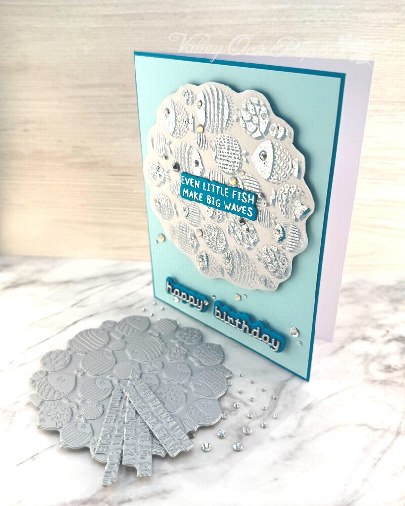 A handmade birthday card with a light turquoise background featuring a scalloped vellum circle filled with silver foiled fish designs in various patterns and textures. In the center of the fish pattern is a dark turquoise sentiment with silver text reading "EVEN LITTLE FISH MAKE BIG WAVES." 

Below the circular design, "happy birthday" appears in silver cursive lettering on a dark turquoise shadow layer. Small clear rhinestones or gems are scattered across the card for decoration. 

The card is photographed standing on a white marble surface with BetterPress plates with fish designs and a few sentiments are visible in the bottom left corner.