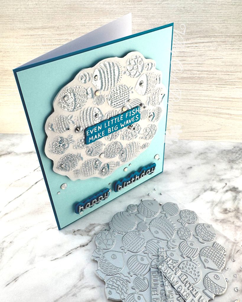 A handmade birthday card with a light turquoise background featuring a scalloped vellum circle filled with silver foiled fish designs in various patterns and textures. In the center of the fish pattern is a dark turquoise sentiment with silver text reading "EVEN LITTLE FISH MAKE BIG WAVES." 

Below the circular design, "happy birthday" appears in silver cursive lettering on a dark turquoise shadow layer. Small clear rhinestones or gems are scattered across the card for decoration. 

The card is photographed standing on a white marble surface with BetterPress plates with fish designs and a few sentiments are visible in the bottom right corner.