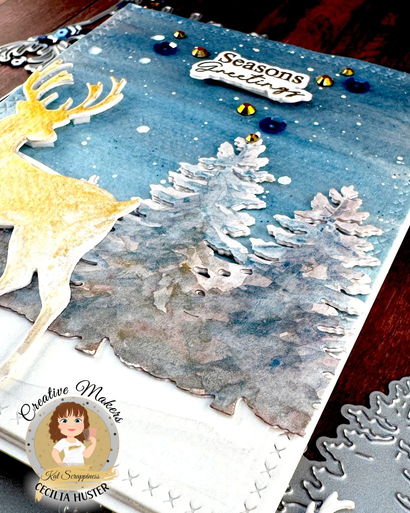 A handmade winter holiday card featuring a layered paper craft design. The card shows a watercolor winter scene with a golden deer silhouette standing in snow, surrounded by white pine trees against a blue starry sky. The text 'Seasons Greetings' appears at the top in gold lettering.
The card is decorated with blue and gold sequins scattered across the surface. It's lying on a dark wooden surface and has been photographed from the bottom right corner.
A circular logo in the bottom left identifies this as work by Kat Scrappiness designer Cecilia Huster, featuring a cartoon avatar of a woman with brown hair.