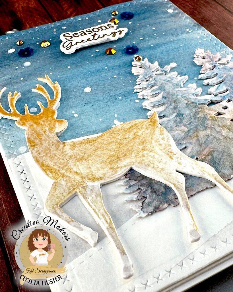 A handmade winter holiday card featuring a layered paper craft design. The card shows a watercolor winter scene with a golden deer silhouette standing in snow, surrounded by white pine trees against a blue starry sky. The text 'Seasons Greetings' appears at the top in gold lettering.
The card is decorated with blue and gold sequins scattered across the surface. It's lying on a dark wooden surface and has been photographed from the bottom left corner.
A circular logo in the bottom left identifies this as work by Kat Scrappiness designer Cecilia Huster, featuring a cartoon avatar of a woman with brown hair.