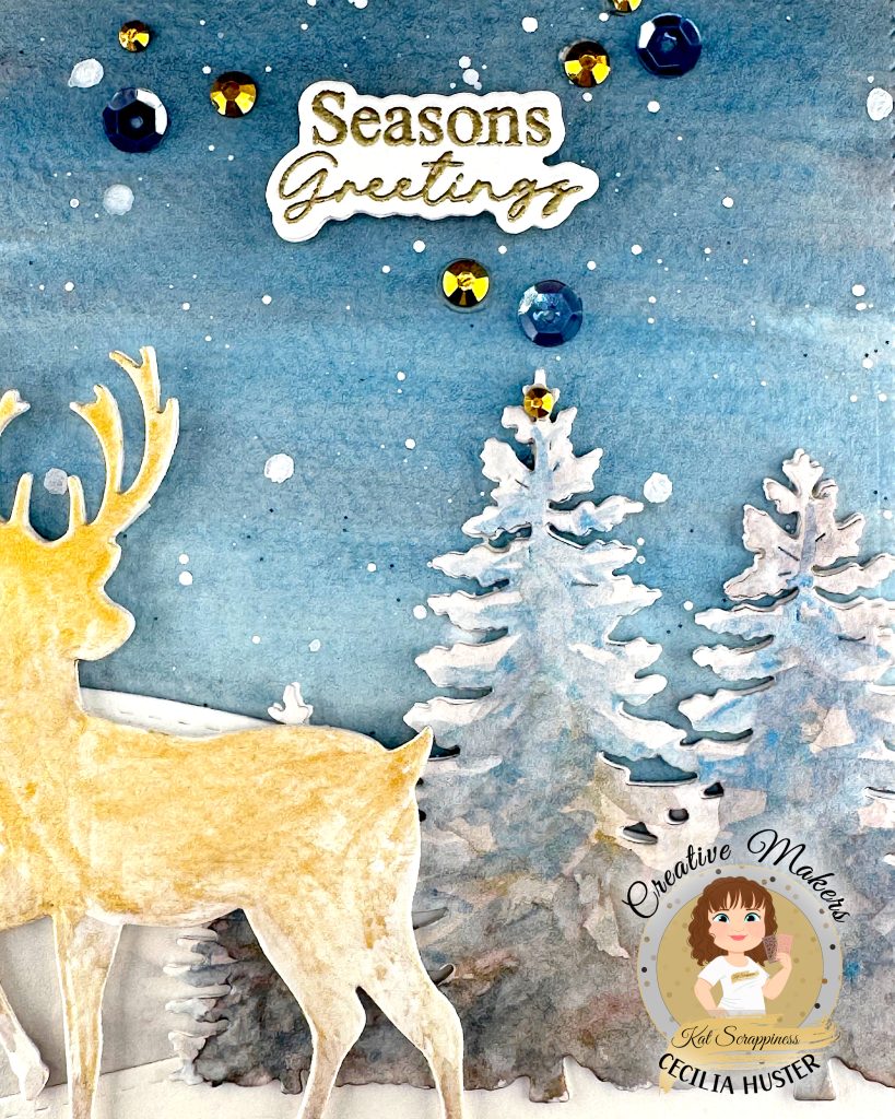 A handmade winter holiday card featuring a layered paper craft design. The card shows a watercolor winter scene with a golden deer silhouette standing in snow, surrounded by white pine trees against a blue starry sky. The text 'Seasons Greetings' appears at the top in gold lettering.
The card is decorated with blue and gold sequins scattered across the surface. This is a close-up showing only parts of the deer and trees and the whole sentiment.
A circular logo in the bottom right identifies this as work by Kat Scrappiness designer Cecilia Huster, featuring a cartoon avatar of a woman with brown hair.