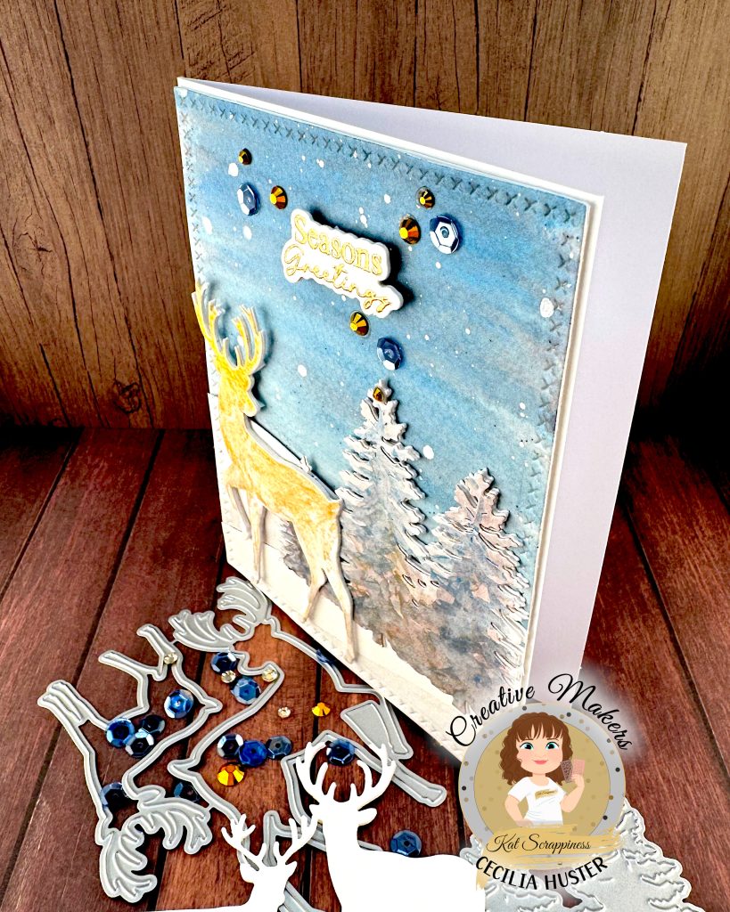 A handmade winter holiday card featuring a layered paper craft design. The card shows a watercolor winter scene with a golden deer silhouette standing in snow, surrounded by white pine trees against a blue starry sky. The text 'Seasons Greetings' appears at the top in gold lettering.
The card is decorated with blue and gold sequins scattered across the surface. It's standing up facing left. In front of it are reindeer die cuts, deer dies, blue sequins and gold rhinestones.
A circular logo in the bottom left identifies this as work by Kat Scrappiness designer Cecilia Huster, featuring a cartoon avatar of a woman with brown hair.