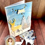 A handmade winter holiday card featuring a layered paper craft design. The card shows a watercolor winter scene with a golden deer silhouette standing in snow, surrounded by white pine trees against a blue starry sky. The text 'Seasons Greetings' appears at the top in gold lettering. The card is decorated with blue and gold sequins scattered across the surface. It's standing up facing right. In front of it are reindeer die cuts, a tree die, blue sequins and gold rhinestones. A circular logo in the bottom left identifies this as work by Kat Scrappiness designer Cecilia Huster, featuring a cartoon avatar of a woman with brown hair.