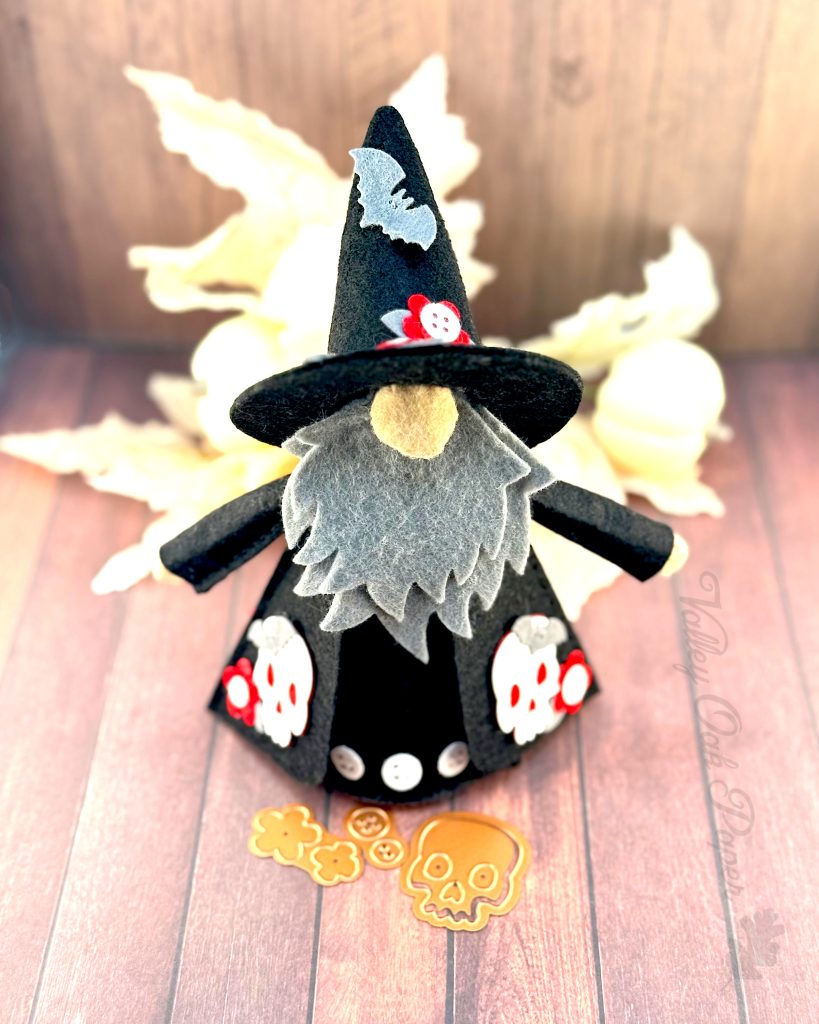 A handmade felt Halloween gnome with a gray beard is seen from the front. The gnome is wearing a black pointed witchy hat adorned with a bat silhouette and small red and flowers, and a black robe with more red flower patches. 

The gnome stands on weathered wooden planks with white pumpkins and leaves in the background. Halloween-themed dies (flowers, buttons, and a skull) are scattered in front.