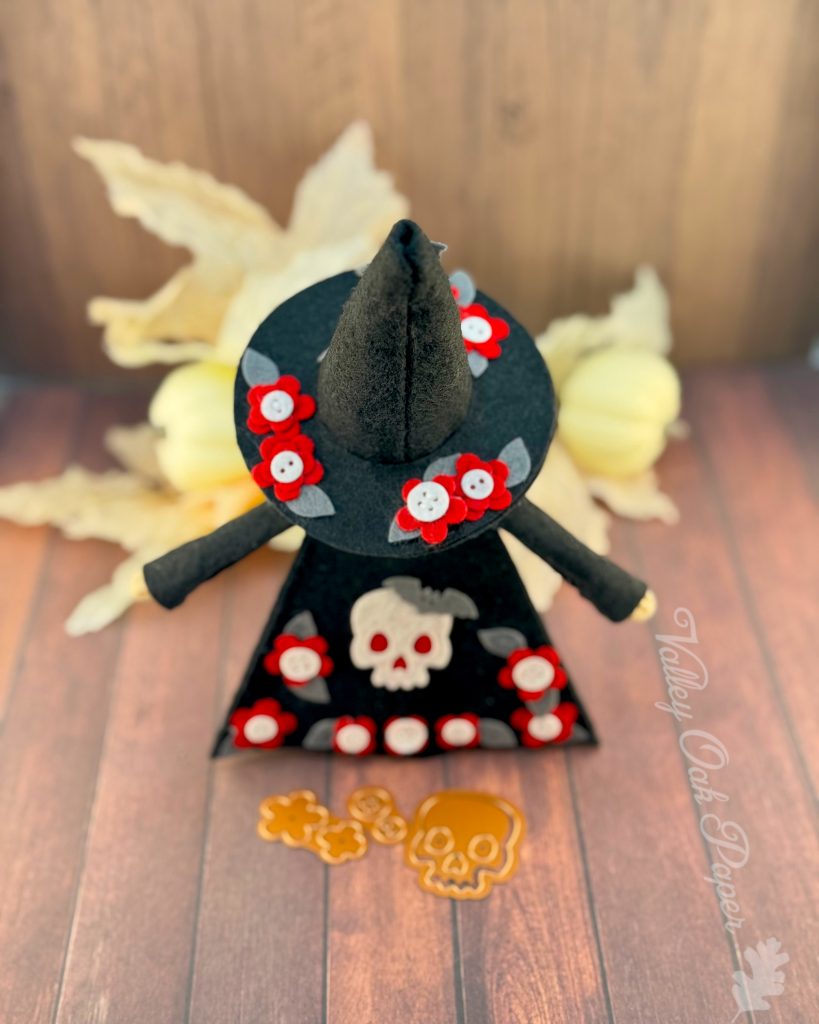 A handmade felt Halloween gnome with a gray beard is seen from behind. The gnome is wearing a black pointed witchy hat adorned with small red flowers, and a black robe with a big white skull and more red flower patches. 

The gnome stands on weathered wooden planks with white pumpkins and leaves in the background. Halloween-themed dies (flowers, buttons, and a skull) are scattered in front.
