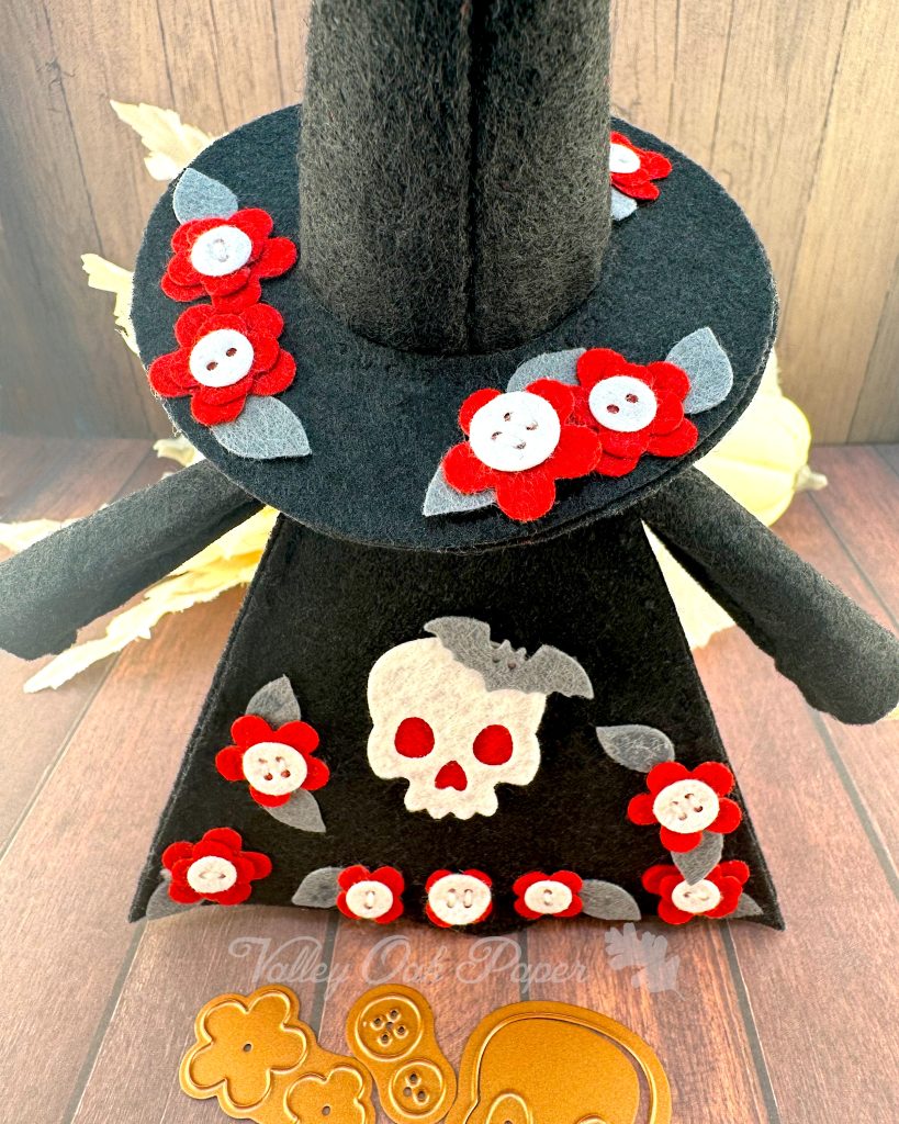 A handmade felt Halloween gnome with a gray beard is seen from behind. The gnome is wearing a black pointed witchy hat adorned with small red flowers. In this photo you can clearly see that the flower centers are buttons. They are also wearing a black robe with a big white skull and more red flower patches. 

The gnome stands on weathered wooden planks with white pumpkins and leaves in the background. Halloween-themed dies (flowers, buttons, and a skull) are scattered in front.