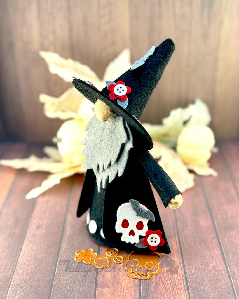 A handmade felt Halloween gnome with a gray beard is seen in profile. The gnome is wearing a black pointed witchy hat adorned with small red flowers, and a black robe with a big white skull and more red flower patches. 

The gnome stands on weathered wooden planks with white pumpkins and leaves in the background. Halloween-themed dies (flowers, buttons, and a skull) are scattered in front.