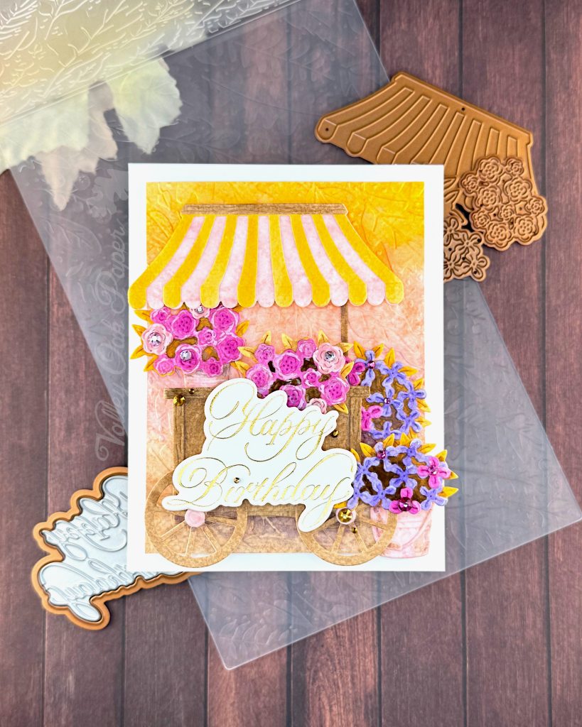 A handmade birthday card featuring a charming flower cart design sits on a wooden surface.
The card shows a yellow and pink striped awning over a wooden cart overflowing with pink roses and purple blooms. It has an embossed background in yellow, pink and brown. The gold foil copperplate greeting "Happy Birthday" is prominently displayed on the cart.
Around the card are crafting supplies including ornate metal dies - one appears to be an awning design and another shows floral patterns. An embossing folder is also visible.