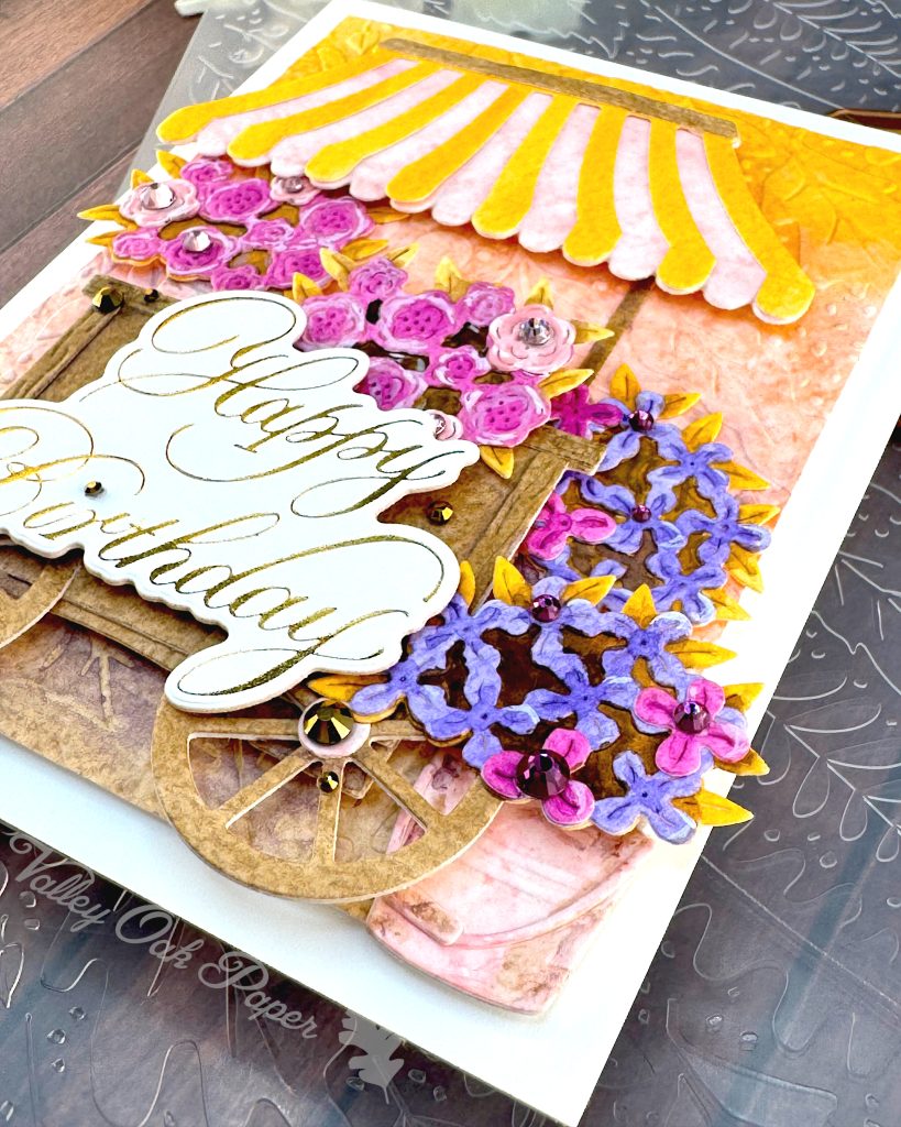A handmade birthday card featuring a charming flower cart design sits on a wooden surface.
The card shows a yellow and pink striped awning over a wooden cart overflowing with pink roses and purple blooms. It has an embossed background in yellow, pink and brown. The gold foil copperplate greeting "Happy Birthday" is prominently displayed on the cart. The photo is taken with the bottom right corner in focus. A wheel is visible with a gold gem in the center of it. Behind the wheel sits a pink bucket overflowing with purple flowers.
An embossing folder is visible in the background.