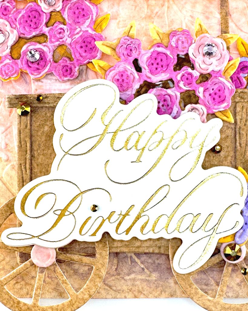A closeup of a handmade birthday card.
The closeup shows a wooden cart with a prominent "Happy Birthday" greeting. The cart appears to contain many pink roses. Some of the roses have pink jewels in their centers. A few gold gems are scattered around and on the sentiment.