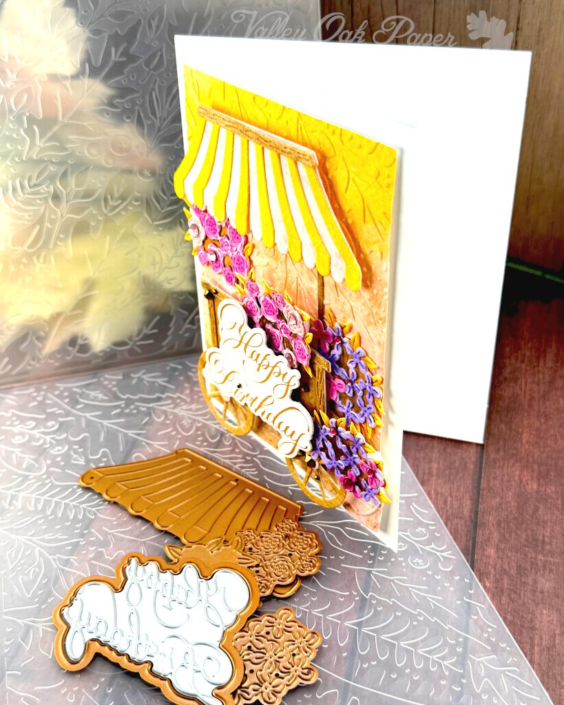 A handmade birthday card featuring a charming flower cart design stands on a wooden surface. The card is facing left.
The card shows a yellow and pink striped awning over a wooden cart overflowing with pink roses and purple blooms. It has an embossed background in yellow, pink and brown. The gold foil copperplate greeting "Happy Birthday" is prominently displayed on the cart.
Around the card are crafting supplies including ornate metal dies - one appears to be an awning design and another shows floral patterns. An embossing folder is also visible in the background.