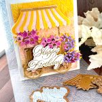 A handmade birthday card featuring a charming flower cart design stands on a wooden surface. The card shows a yellow and pink striped awning over a wooden cart overflowing with pink roses and purple blooms. It has an embossed background in yellow, pink and brown. The gold foil copperplate greeting "Happy Birthday" is prominently displayed on the cart. Around the card are crafting supplies including ornate metal dies - one appears to be an awning design and another shows floral patterns. An embossing folder is also visible in the background.