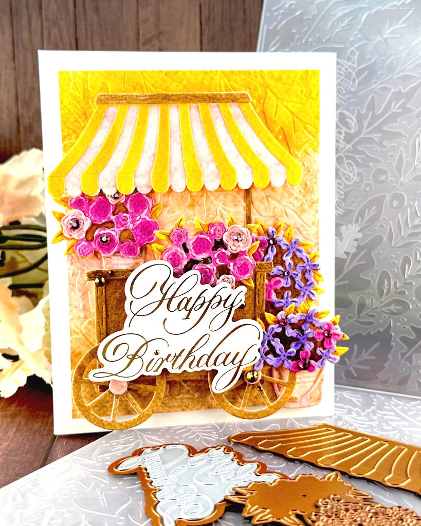 A handmade birthday card featuring a charming flower cart design stands on a wooden surface. The card is facing right.
The card shows a yellow and pink striped awning over a wooden cart overflowing with pink roses and purple blooms. It has an embossed background in yellow, pink and brown. The gold foil copperplate greeting "Happy Birthday" is prominently displayed on the cart.
Around the card are crafting supplies including ornate metal dies - one appears to be an awning design and another shows floral patterns. An embossing folder is also visible in the background.