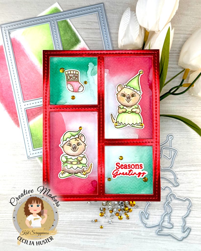 Handmade holiday greeting card featuring a red frame with four panels containing watercolor backgrounds in red and green. The card displays two cute quokka characters wearing green elf costumes, along with a Christmas stocking design. One panel contains the text 'Seasons Greetings' in red script. The card is decorated with gold sequins and sits on a white wooden surface alongside white flowers, gold rhinestones and some of the dies used to create it. A circular logo in the bottom left says 'Kat Scrappiness Creative Maker Cecilia Huster.'