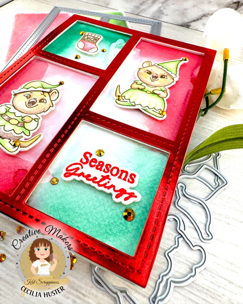 Handmade holiday greeting card featuring a red frame with four panels containing watercolor backgrounds in red and green. The card displays two cute quokka characters wearing green elf costumes, along with a Christmas stocking design. 

Closest to the observer,  a panel contains the text 'Seasons Greetings' in red script. The card is decorated with gold rhinestones and sits on a white wooden surface alongside white flowers, gold rhinestones and some of the dies used to create it. A circular logo in the bottom left says 'Kat Scrappiness Creative Maker Cecilia Huster.'