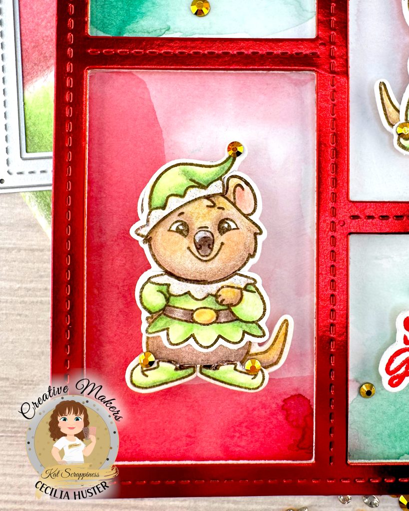 Closeup of a holiday greeting card featuring a red frame. The focus is on a rectangular opening in the frame with a red watercolor background. A cute boy quokka wearing a green elf costume is centered in the opening. Gold rhinestones adorn her elf slippers and hat. The card sits on a white wooden surface alongside craft supplies.

A circular logo in the bottom left says 'Kat Scrappiness Creative Maker Cecilia Huster.'