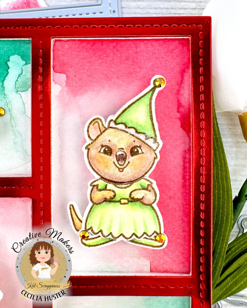 Closeup of a holiday greeting card featuring a red frame. The focus is on a rectangular opening in the frame with a red watercolor background. A cute girl quokka wearing a green elf costumeis centered in the opening. Gold rhinestones adorn her elf slippers and hat. The card sits on a white wooden surface alongside white flowers.

A circular logo in the bottom left says 'Kat Scrappiness Creative Maker Cecilia Huster.'