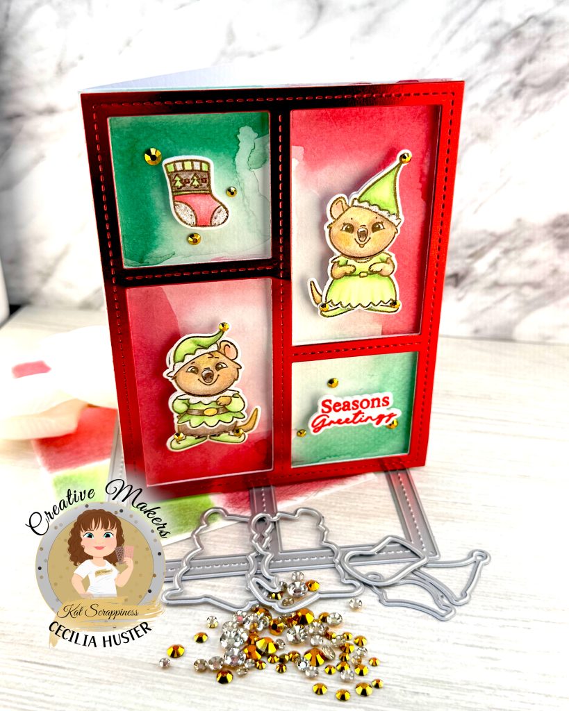 Handmade holiday greeting card featuring a red frame with four panels containing watercolor backgrounds in red and green. The card displays two cute quokka characters wearing green elf costumes, along with a Christmas stocking design. One panel contains the text 'Seasons Greetings' in red script. The card is decorated with gold sequins and stands facing right on a white wooden surface alongside white flowers, gold rhinestones and some of the dies used to create it. A circular logo in the bottom left says 'Kat Scrappiness Creative Maker Cecilia Huster.'