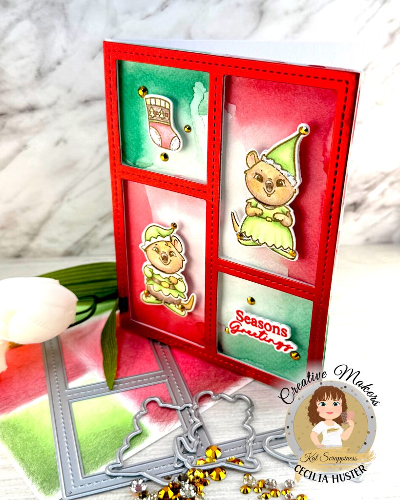 Handmade holiday greeting card featuring a red frame with four panels containing watercolor backgrounds in red and green. The card displays two cute quokka characters wearing green elf costumes, along with a Christmas stocking design. One panel contains the text 'Seasons Greetings' in red script. The card is decorated with gold sequins and stands facing left on a white wooden surface alongside white flowers, gold rhinestones and some of the dies used to create it. A circular logo in the bottom left says 'Kat Scrappiness Creative Maker Cecilia Huster.'