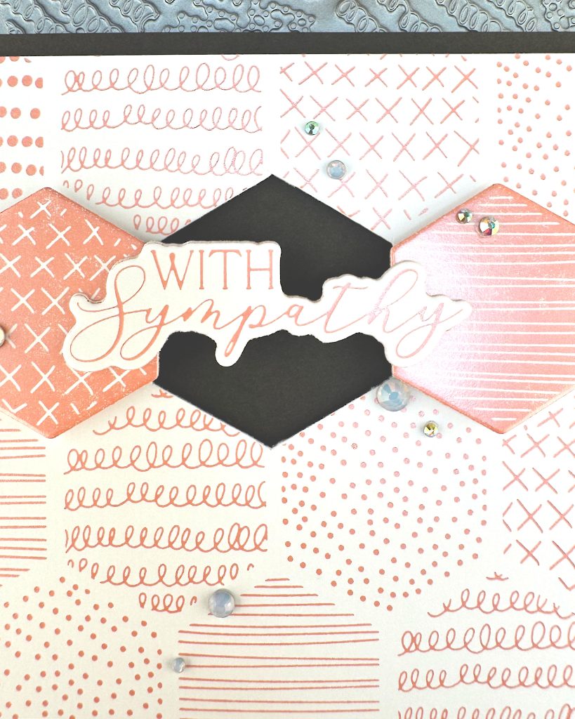A closeup of a handmade greeting card. The card features a soft peach and white patterned background with various decorative elements including dots, crosses, and lined patterns. It is matted in black.

In the center two hexagonal tiles flank a black hexagonal void. The void is bridged by the greeting "With Sympathy." Small rhinestone embellishments are scattered across the card. 