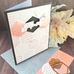 A handmade greeting card sits on a wooden surface surrounded by crafting materials. The card features a soft peach and white patterned background with various decorative elements including dots, crosses, and lined patterns. It is matted in black. In the center two hexagonal tiles flank a black hexagonal void. The void is bridged by the greeting "With Sympathy." Small rhinestone embellishments are scattered across the card. The card is standing on crafting supplies including the press plate used to create it. Autumn leaves in warm orange and yellow hues lie behind it. All items are arranged on a dark wood grain background.