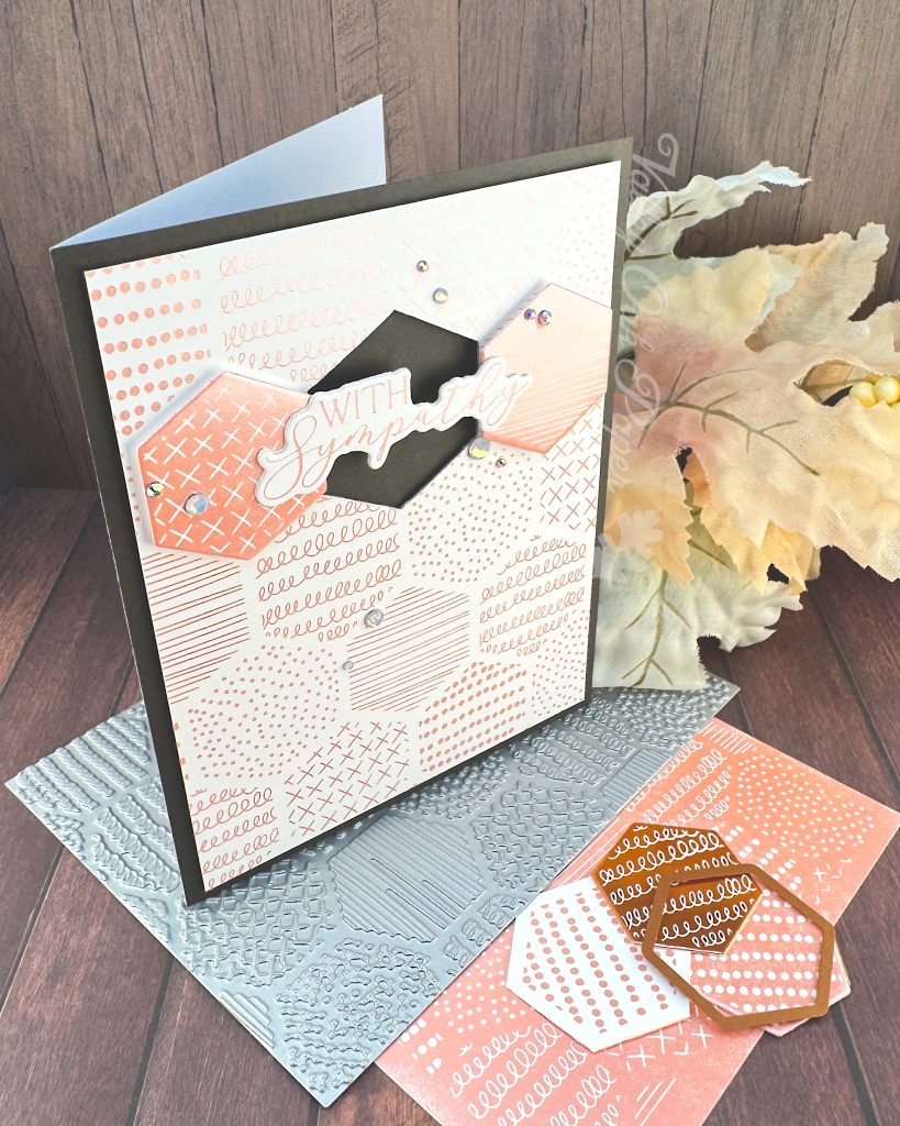 A handmade greeting card sits on a wooden surface surrounded by crafting materials. The card features a soft peach and white patterned background with various decorative elements including dots, crosses, and lined patterns. It is matted in black.

In the center two hexagonal tiles flank a black hexagonal void. The void is bridged by the greeting "With Sympathy." Small rhinestone embellishments are scattered across the card. The card is standing on crafting supplies including the press plate used to create it. Autumn leaves in warm orange and yellow hues lie behind it. All items are arranged on a dark wood grain background.
