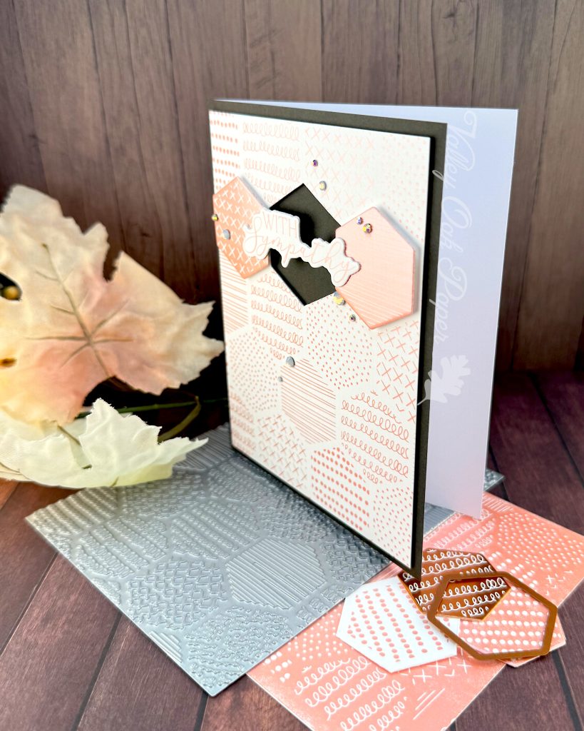 A handmade greeting card sits on a wooden surface surrounded by crafting materials. The card features a soft peach and white patterned background with various decorative elements including dots, crosses, and lined patterns. It is matted in black.

In the center two hexagonal tiles flank a black hexagonal void. The void is bridged by the greeting "With Sympathy." Small rhinestone embellishments are scattered across the card. The card is standing on crafting supplies including the press plate used to create it and autumn leaves in warm orange and yellow hues. All items are arranged on a dark wood grain background.