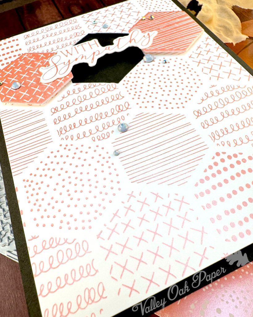 A handmade greeting card sits on a wooden surface surrounded by crafting materials. The card features a soft peach and white patterned background with various decorative elements including dots, crosses, and lined patterns. It is matted in black.

In the center two hexagonal tiles flank a black hexagonal void. The void is bridged by the greeting "With Sympathy." Small rhinestone embellishments are scattered across the card. The card is lying on top of foiled cardstock.  The photo was taken from the bottom left corner of the card. All items are arranged on a dark wood grain background.