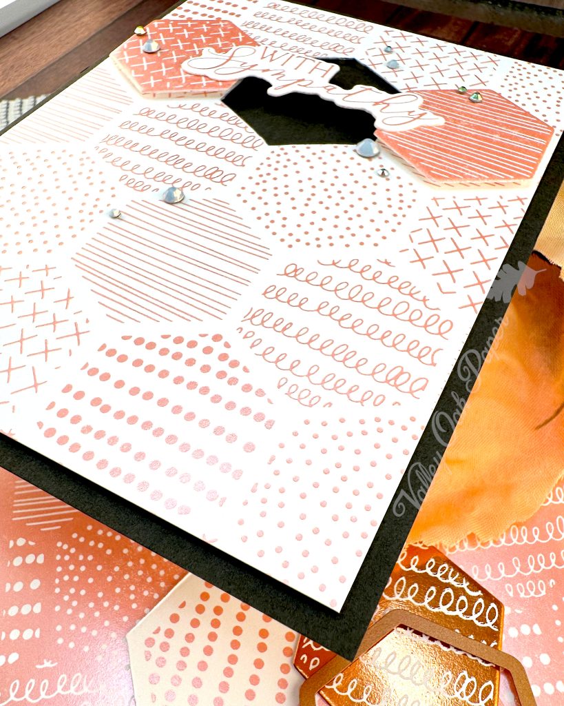 A handmade greeting card sits on a wooden surface surrounded by crafting materials. The card features a soft peach and white patterned background with various decorative elements including dots, crosses, and lined patterns. It is matted in black.

In the center two hexagonal tiles flank a black hexagonal void. The void is bridged by the greeting "With Sympathy." Small rhinestone embellishments are scattered across the card. The card is lying on top of foiled cardstock.  All items are arranged on a dark wood grain background.