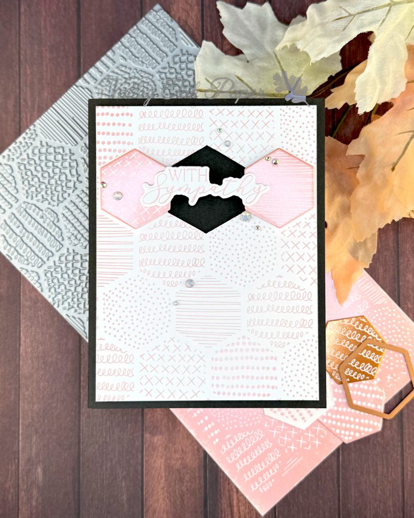 A handmade greeting card sits on a wooden surface surrounded by crafting materials. The card features a soft peach and white patterned background with various decorative elements including dots, crosses, and lined patterns. It is matted in black.

In the center two hexagonal tiles flank a black hexagonal void. The void is bridged by the greeting "With Sympathy." Small rhinestone embellishments are scattered across the card. The card is displayed in front of crafting supplies including the press plate used to create it and autumn leaves in warm orange and yellow hues. All items are arranged on a dark wood grain background.
