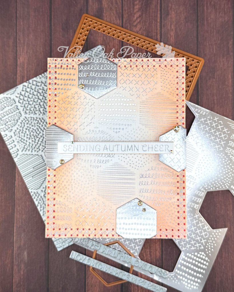 A handmade autumn-themed greeting card displayed on a dark wood surface. The card features a warm peach background with silver foil geometric patterns including dots, crosses, and lines in hexagonal sections. A white banner across the center reads 'SENDING AUTUMN CHEER' in silver lettering. 

The card is layered with hexagonal elements with negative versions of the patterns in the background. Behind the main card lie the BetterPress plates and dies used to create it, as well as  coordinating paper pieces with similar geometric patterns in silver and white. Small gold gems accent some of the layered elements. 

The overall design has a sophisticated autumn color palette of coral, gray, and white with intricate patterned details throughout.