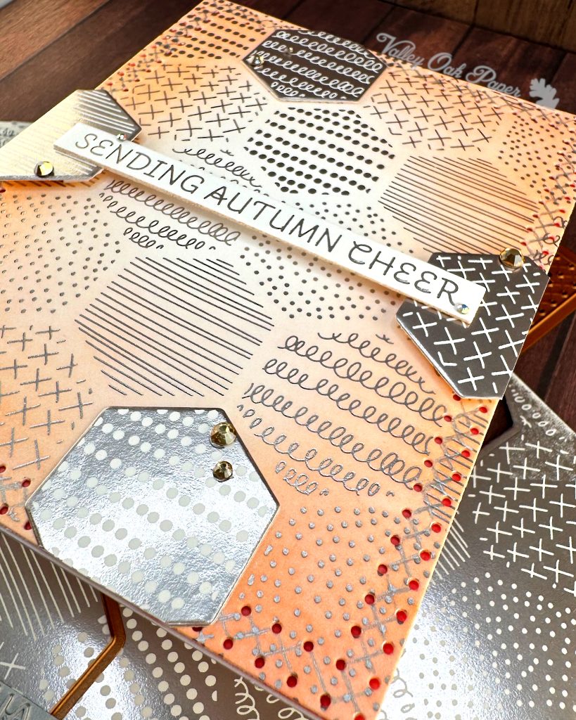 A handmade autumn-themed greeting card is seen from the bottom right corner. The card features a warm peach background with silver foil geometric patterns including dots, crosses, and lines in hexagonal sections. A white banner is suspended across the center. It reads 'SENDING AUTUMN CHEER' in silver lettering. 

The card is layered with hexagonal elements with negative versions of the patterns in the background. Behind the main card lie the BetterPress plates and dies used to create it, as well as the  coordinating paper pieces with similar geometric patterns in silver and white. Small gold gems accent some of the layered elements. 