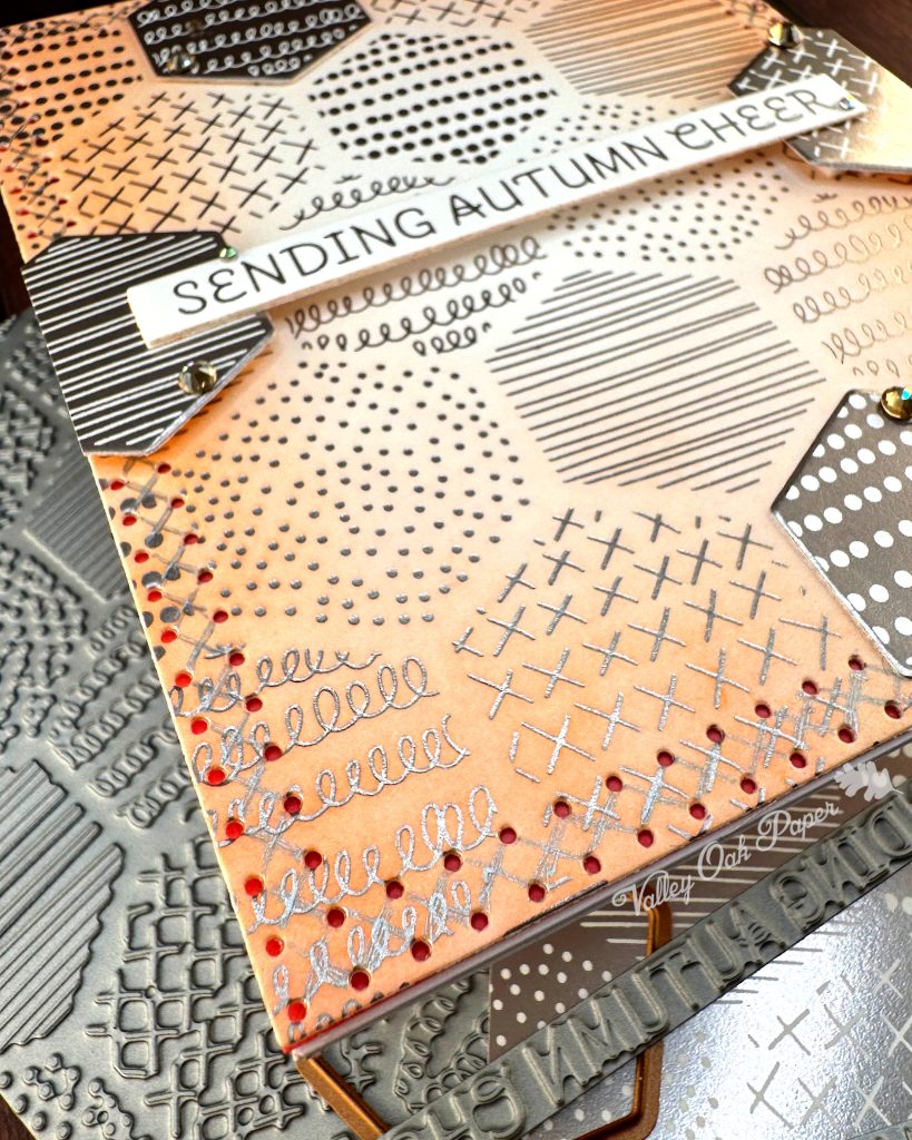 A handmade autumn-themed greeting card is seen from the bottom left corner. The card features a warm peach background with silver foil geometric patterns including dots, crosses, and lines in hexagonal sections. A white banner is suspended across the center. It reads 'SENDING AUTUMN CHEER' in silver lettering. 

The card is layered with hexagonal elements with negative versions of the patterns in the background. Behind the main card lie the BetterPress plates and dies used to create it, as well as the  coordinating paper pieces with similar geometric patterns in silver and white. Small gold gems accent some of the layered elements. 