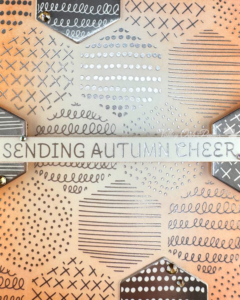 A closeup of a handmade autumn-themed greeting card. The card features a warm peach background with silver foil geometric patterns including dots, crosses, and lines in hexagonal sections. A white banner across the center reads 'SENDING AUTUMN CHEER' in silver lettering. 

The card is layered with hexagonal elements with negative versions of the patterns in the background.  Small gold gems accent some of the layered elements. 