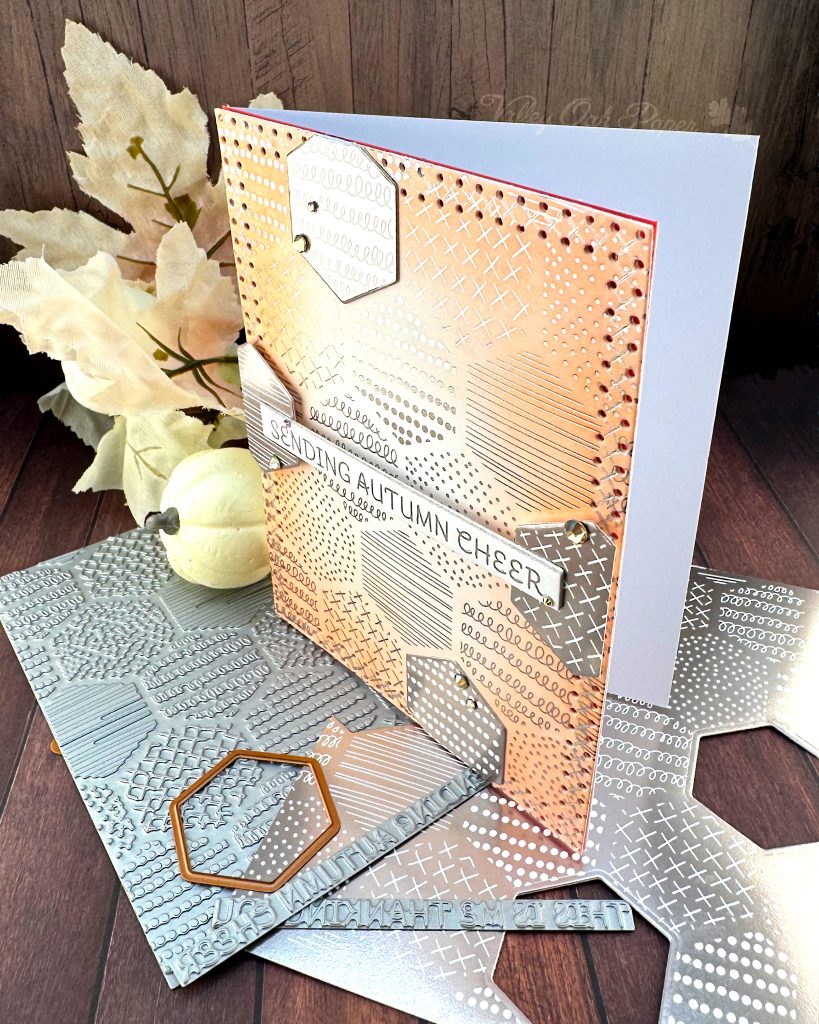 A handmade autumn-themed greeting card stands on a dark wood surface. The card features a warm peach background with silver foil geometric patterns including dots, crosses, and lines in hexagonal sections. A white banner across the center reads 'SENDING AUTUMN CHEER' in silver lettering. 

The card is layered with hexagonal elements with negative versions of the patterns in the background.  Small gold gems accent some of the layered elements. 

The card stands on the BetterPress plates and dies used to create it, as well as  coordinating paper pieces with similar geometric patterns in silver and white. It is facing right. From behind the card some off-white leaves and pumpkins peek through. 

The overall design has a sophisticated autumn color palette of coral, gray, and white with intricate patterned details throughout.