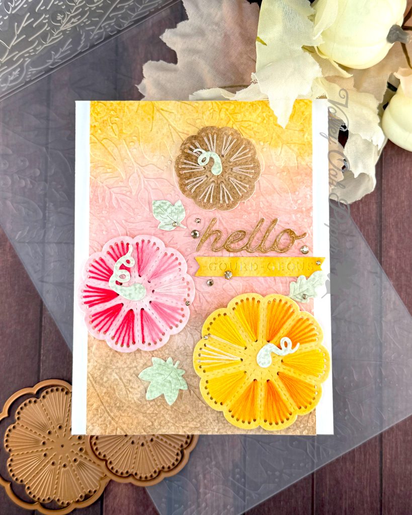 A handmade greeting card with a stitched pumpkin design sits on a wooden surface surrounded by craft supplies. The card features a textured gradient background that transitions from yellow at the top through coral-pink in the middle to brown at the bottom.
Three large pumpkins stitched with embroidery floss are prominently displayed: a small brown pumpkin embroidered with tan floss at the top, a pink pumpkin stitched with variegated red floss on the left, and a large, bright yellow pumpkin stitched with variegated yellow floss at the bottom right.
The word "hello" appears in glossy brown cursive lettering in the center, with a small yellow banner below it. The banner reads "GOURD-EOUS." Small mint green leaf embellishments and gold rhinestone accents are scattered throughout the design.
Under the card an embossing folder with a leaf pattern and a few pumpkin stitching dies are visible. So are some pale pink fabric leaves and mini pumpkins.